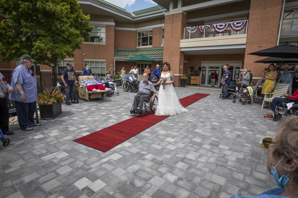A bride walking down a red-carpet aisle at an outdoor wedding ceremony, with wheelchair-using residents watching from both sides